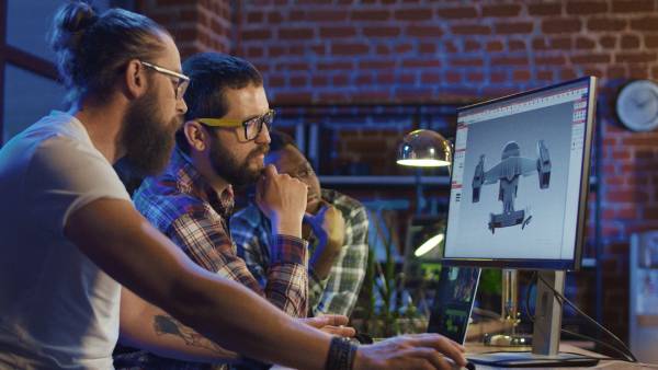 Three men sitting in front of a computer creating and developing graphics for a video game.